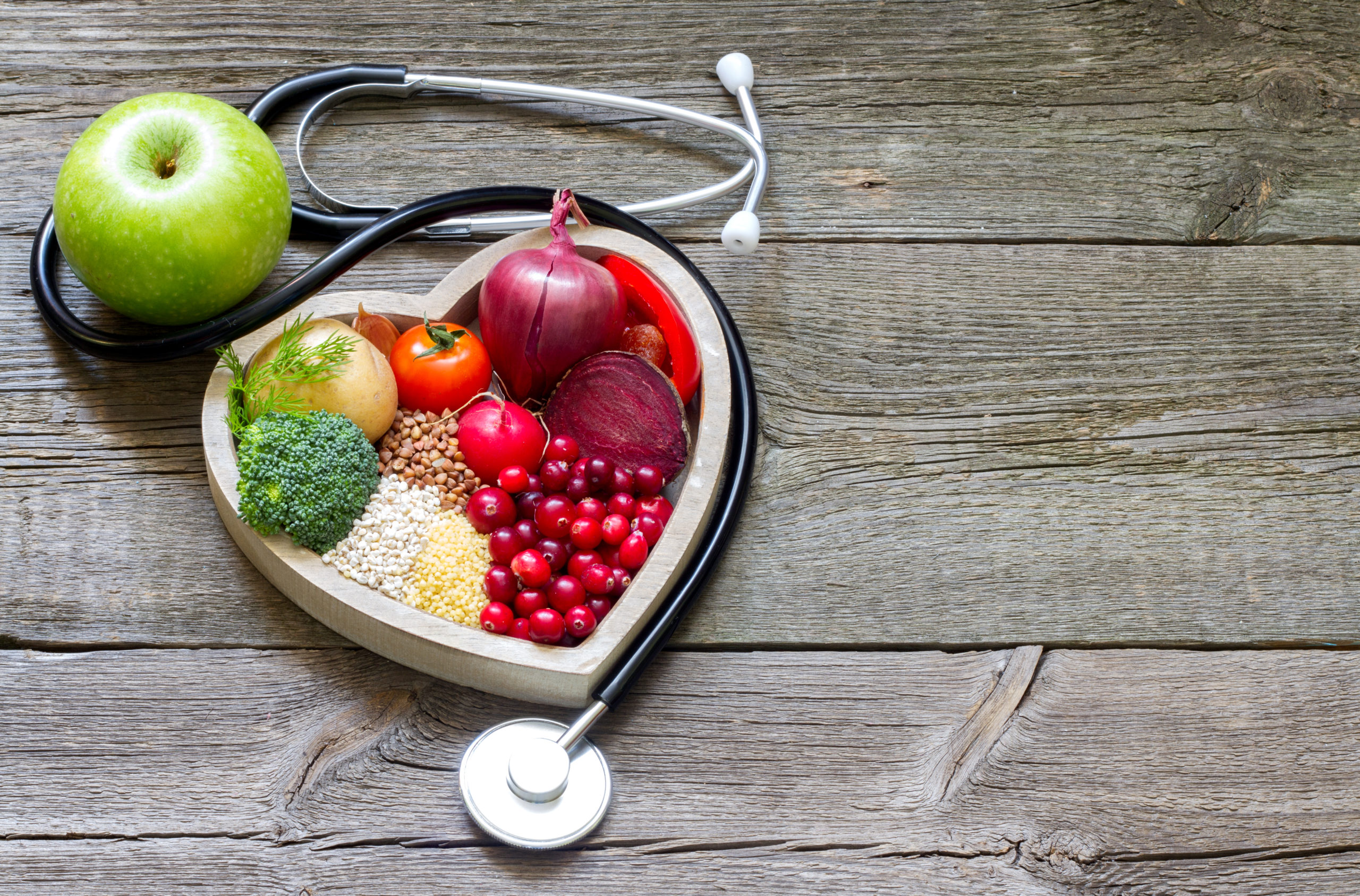photo of a wooden table with fruits and vegetables in a heart shaped bowl with a stethoscope around it